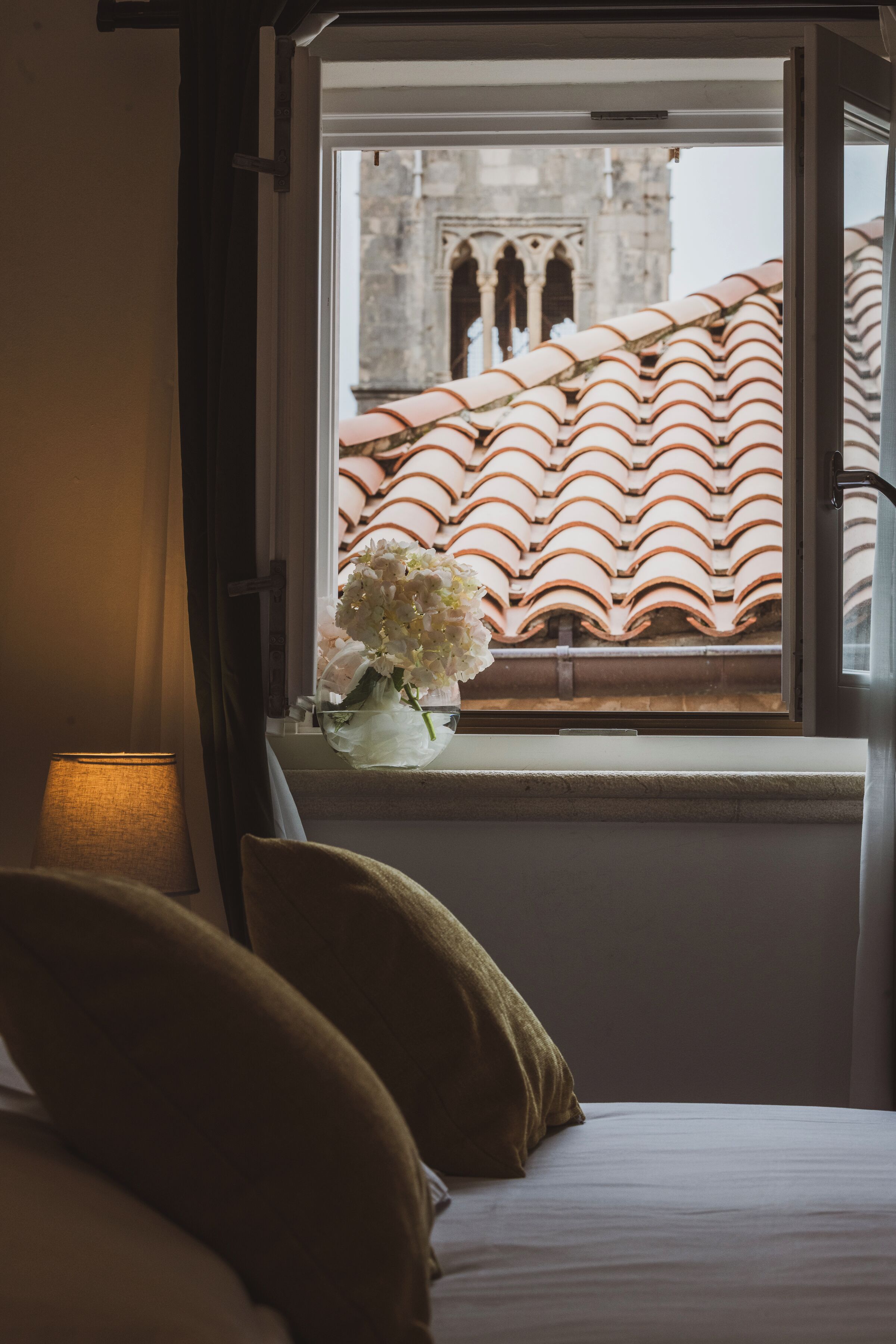 A room with a bed, a window with a view of a building with terracotta roof tiles, and a vase with flowers on the windowsill.