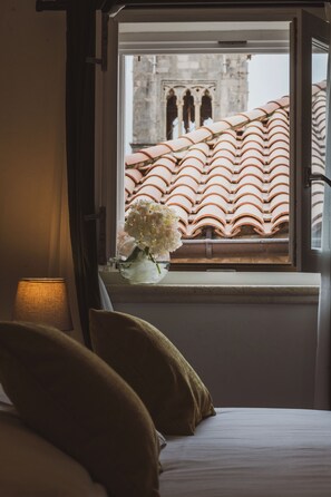 Een kamer met een bed, een raam met uitzicht op een gebouw met terracotta dakpannen, en een bloemenvaas op de vensterbank.