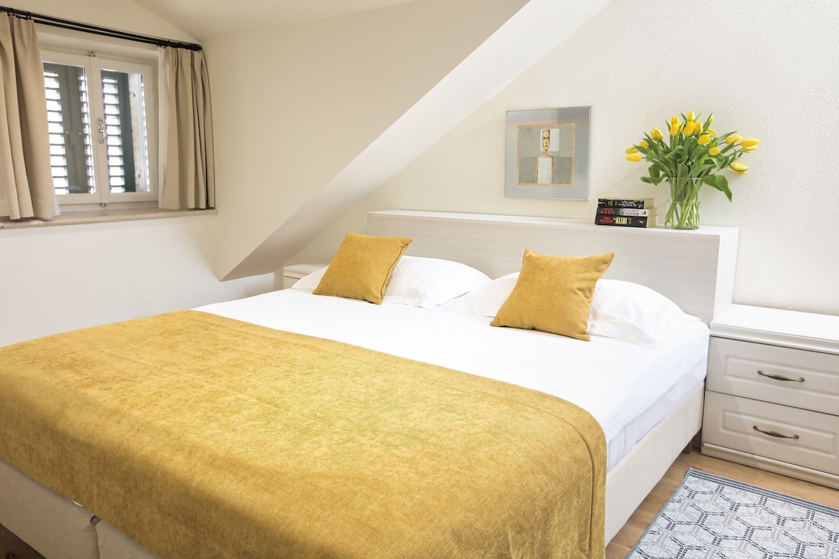 Une chambre à coucher comprenant un lit, une table de chevet, une commode, une fenêtre avec des rideaux, un miroir fixé au mur, un vase avec des fleurs et un tableau encadré.