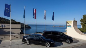 A black car parked in front of a building with flags, including the European Union flag, and a view of the sea.