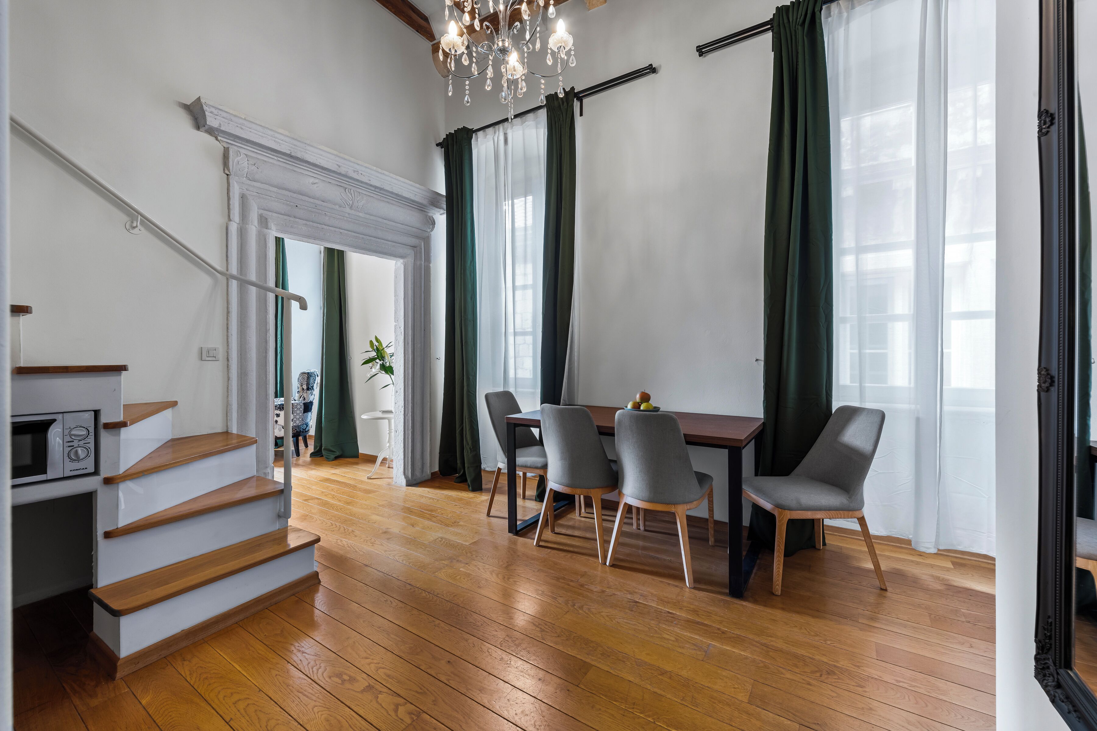 A modern living room with a wooden staircase, a dining area with four chairs, and a chandelier.