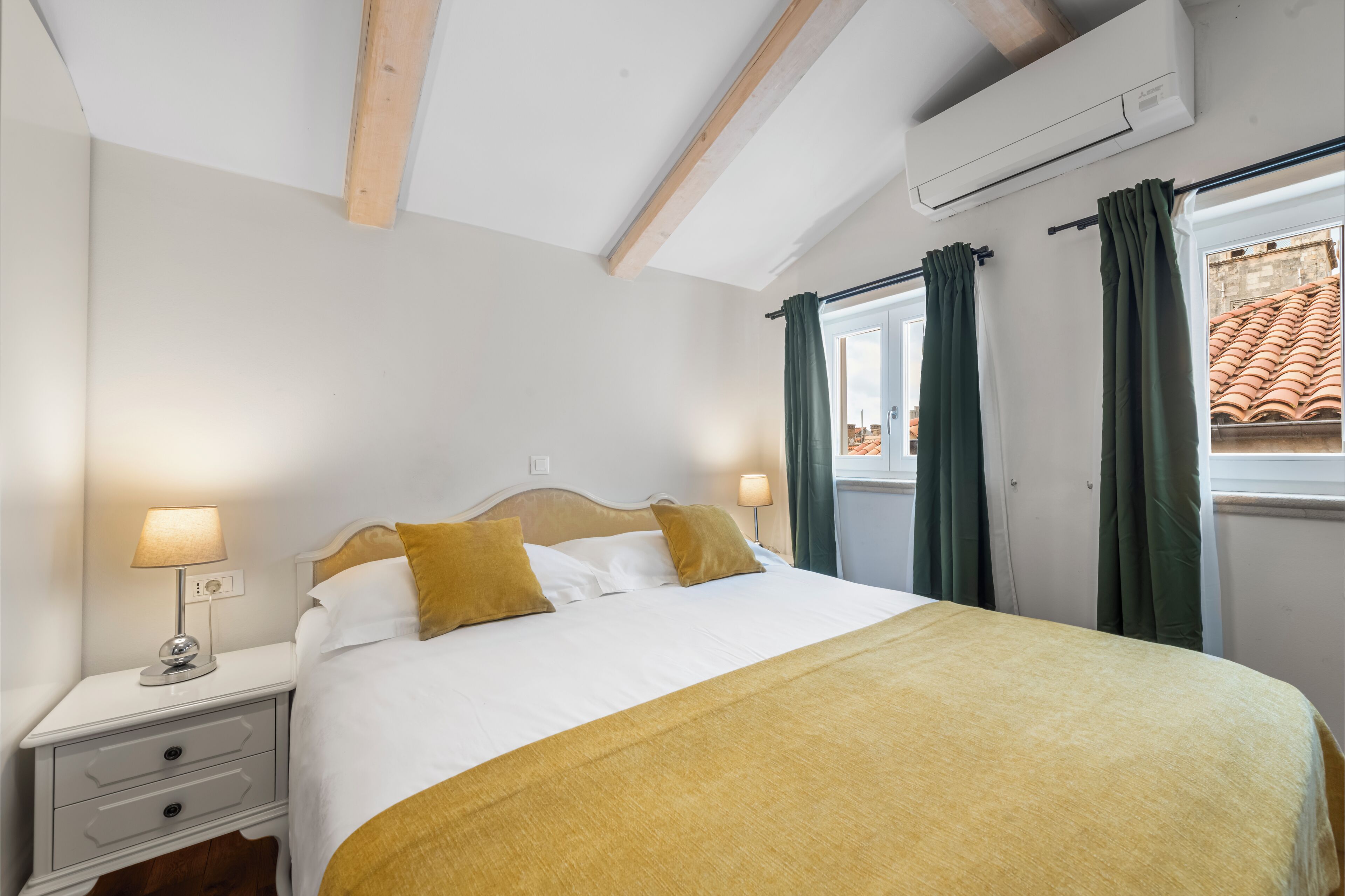 A neatly made bed with a white and yellow bedspread, two pillows, and a bedside lamp. There is a window with green curtains, an air conditioning unit on the wall, and a wooden ceiling beam.