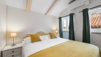 A neatly made bed with a white and yellow bedspread, two pillows, and a bedside lamp. There is a window with green curtains, an air conditioning unit on the wall, and a wooden ceiling beam.