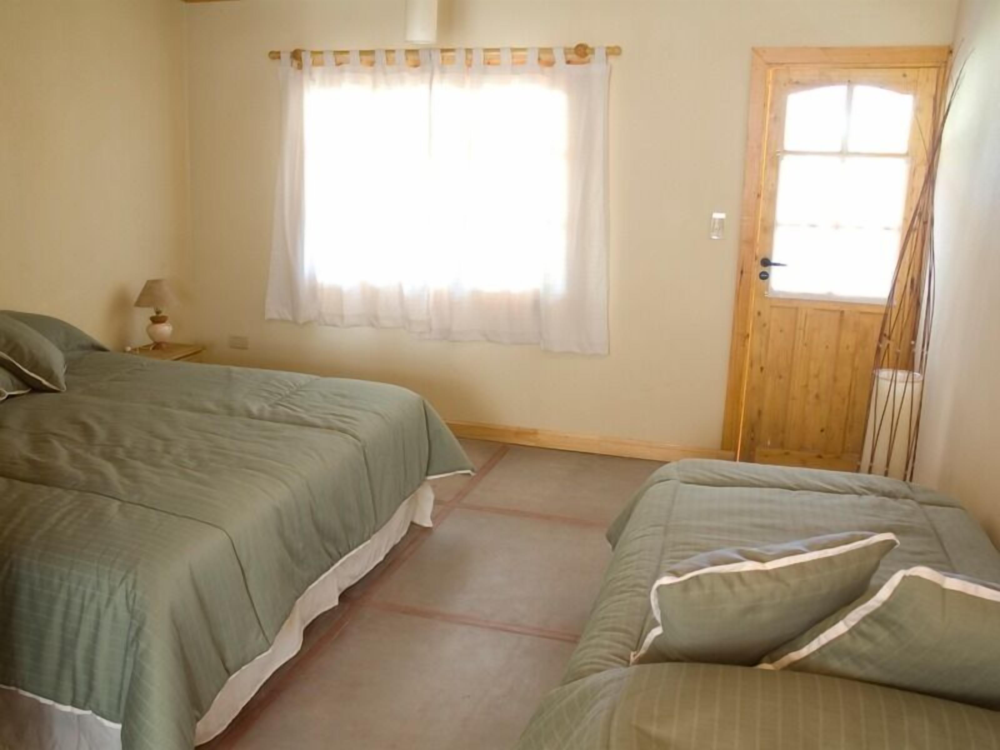 Une chambre avec deux lits, une fenêtre avec des rideaux, une porte en bois et une petite table de chevet avec une lampe.