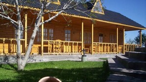 A wooden cabin with a porch, a tree in front, and a flower bed. - Vistalba Lodge (Luján de Cuyo)