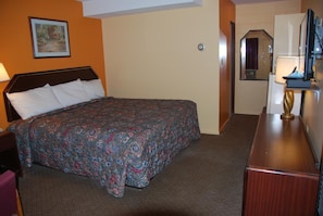 Habitación de hotel con cama, escritorio, silla, lámpara, espejo y un cuadro en la pared.
