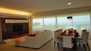 A modern living room with a white sofa, dining table with chairs, and a TV mounted on the wall.