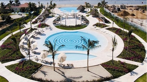 A resort with a central circular pool, lounge areas with sunbeds, and a view of the ocean.