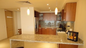 A kitchen with granite countertops, wooden cabinets, a stainless steel refrigerator, a coffee machine, and a bar stool.