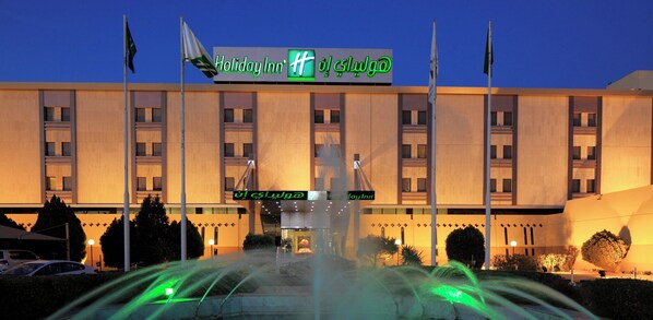 A Holiday Inn hotel with a fountain in front, multiple flags, and a clear sky.
