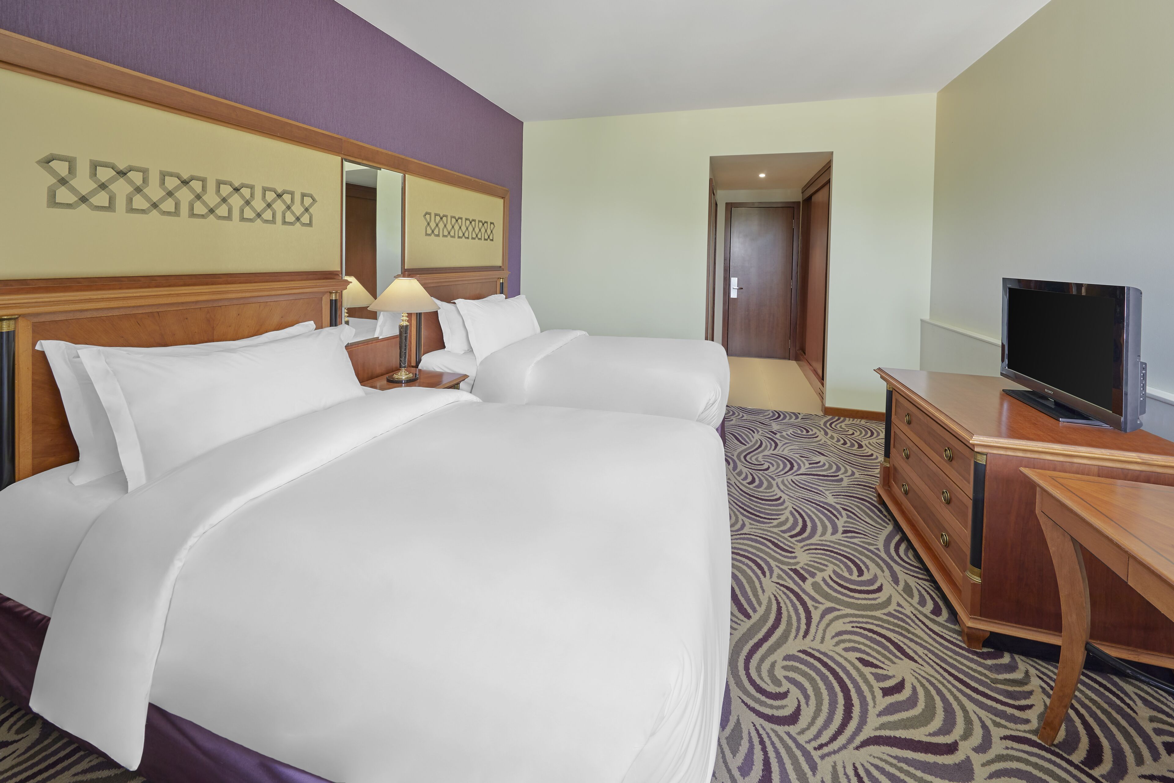 A hotel room with a large bed, a desk, a television, and a patterned carpet.