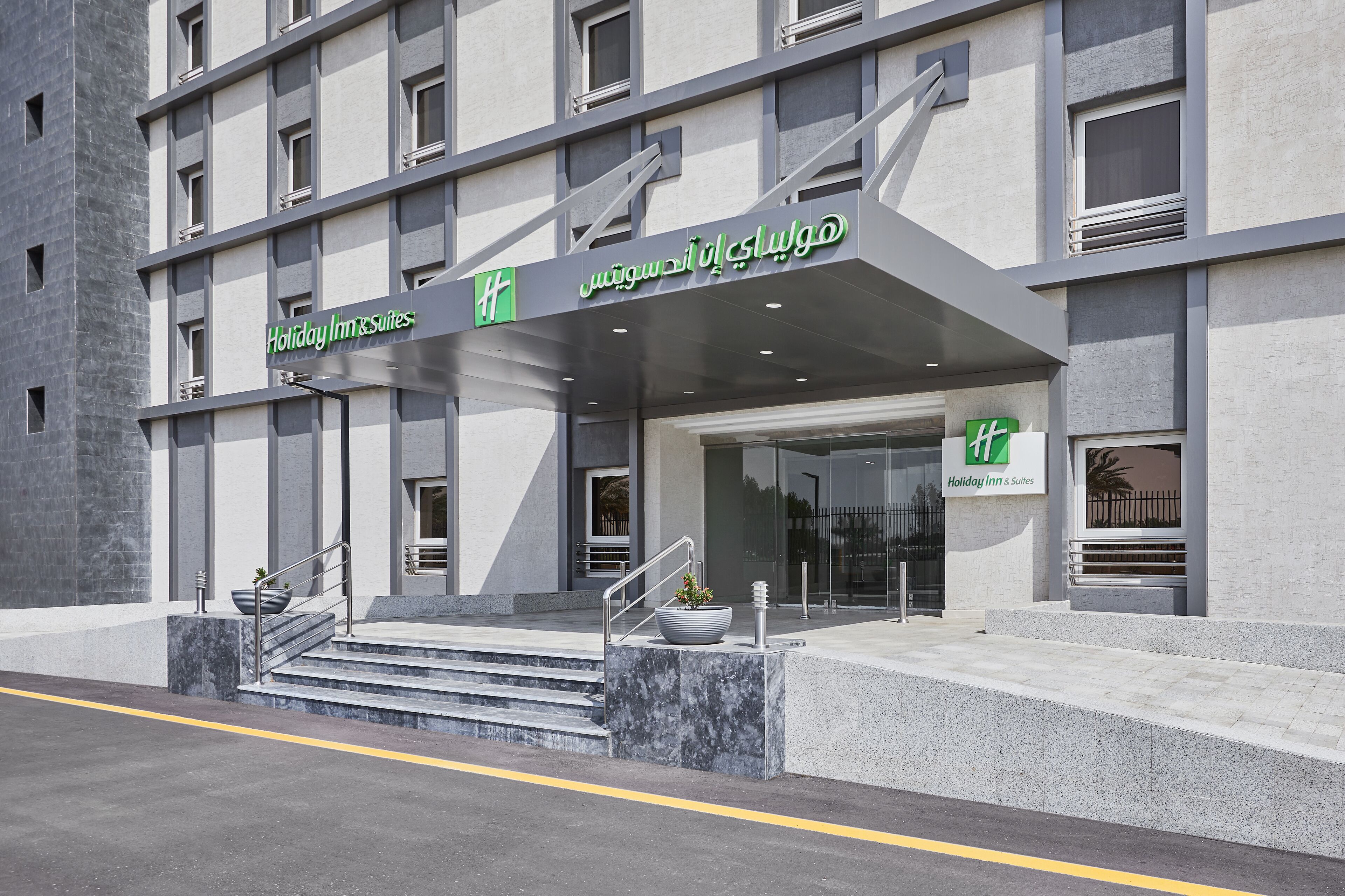 A modern hotel entrance with a staircase leading to the lobby, a large planter with a plant, and a sign indicating the hotel name.