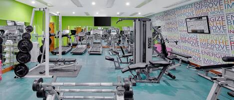 A gym with various exercise equipment, a green wall with motivational words, and a large screen.