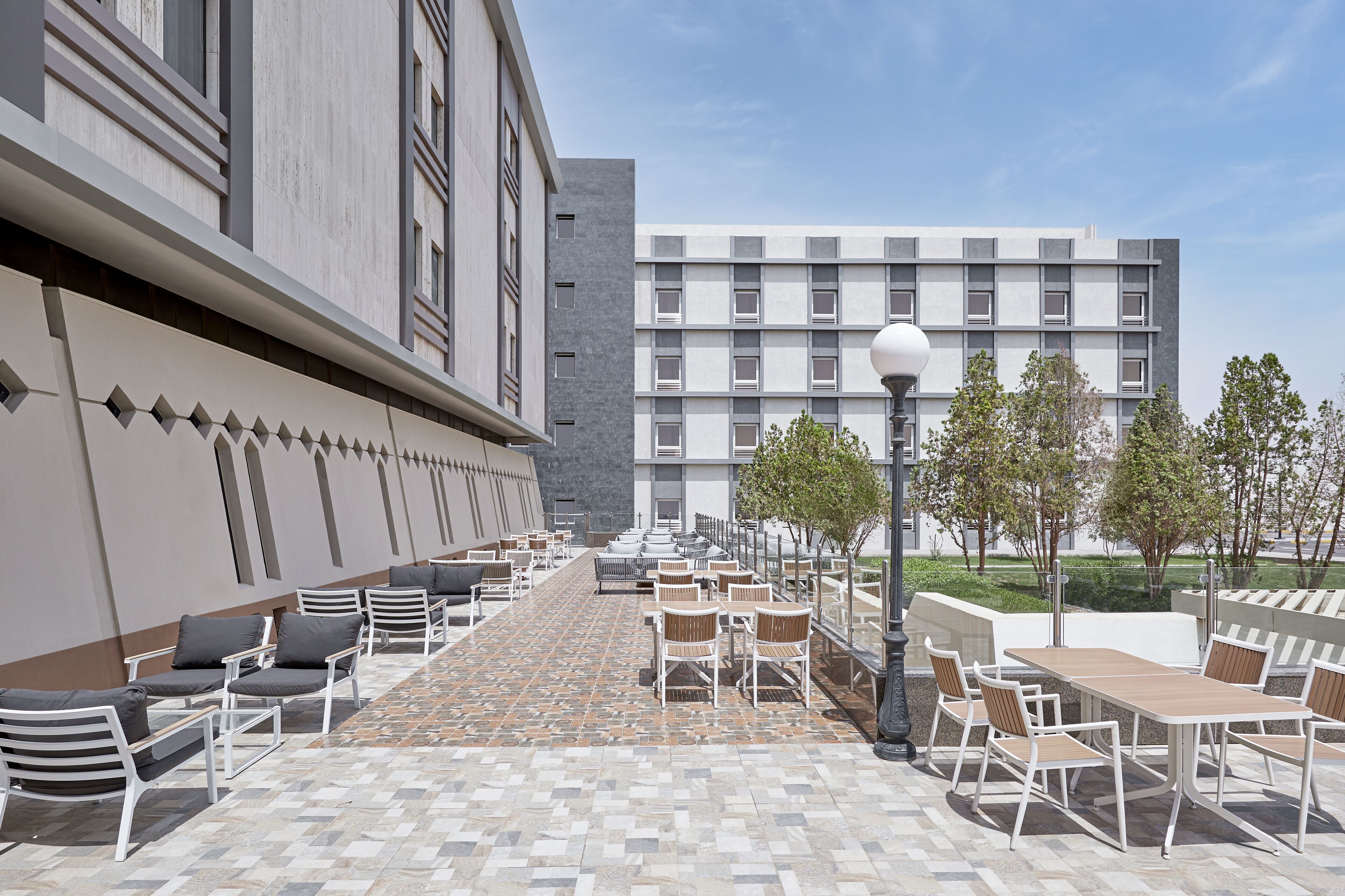 Egy szabadtéri terasz ülőhelyekkel és asztalokkal, a háttérben egy modern épülettel, és tiszta, felhőtlen égbolttal.
