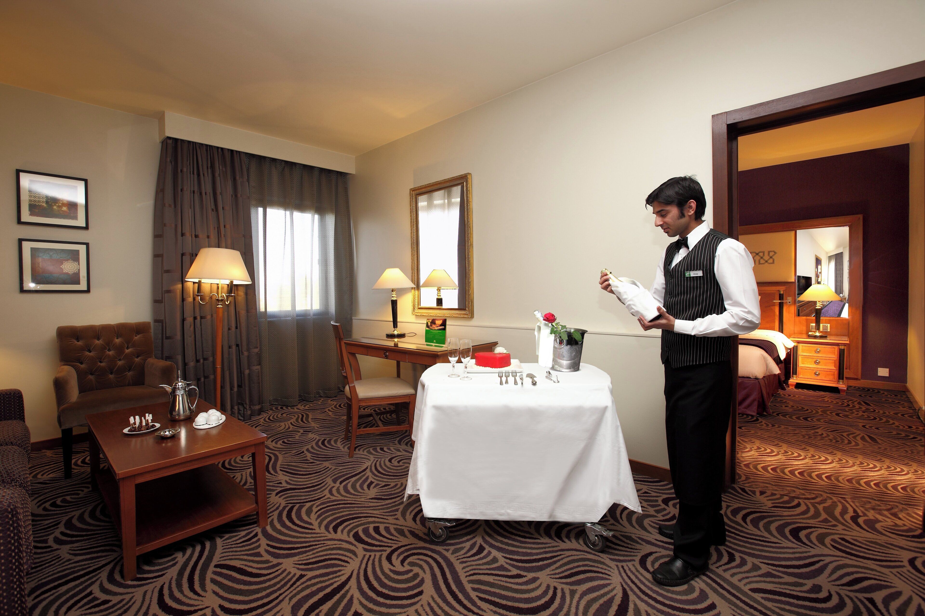 A hotel room with a dining table set, a sofa, a chair, a lamp, and a mirror.