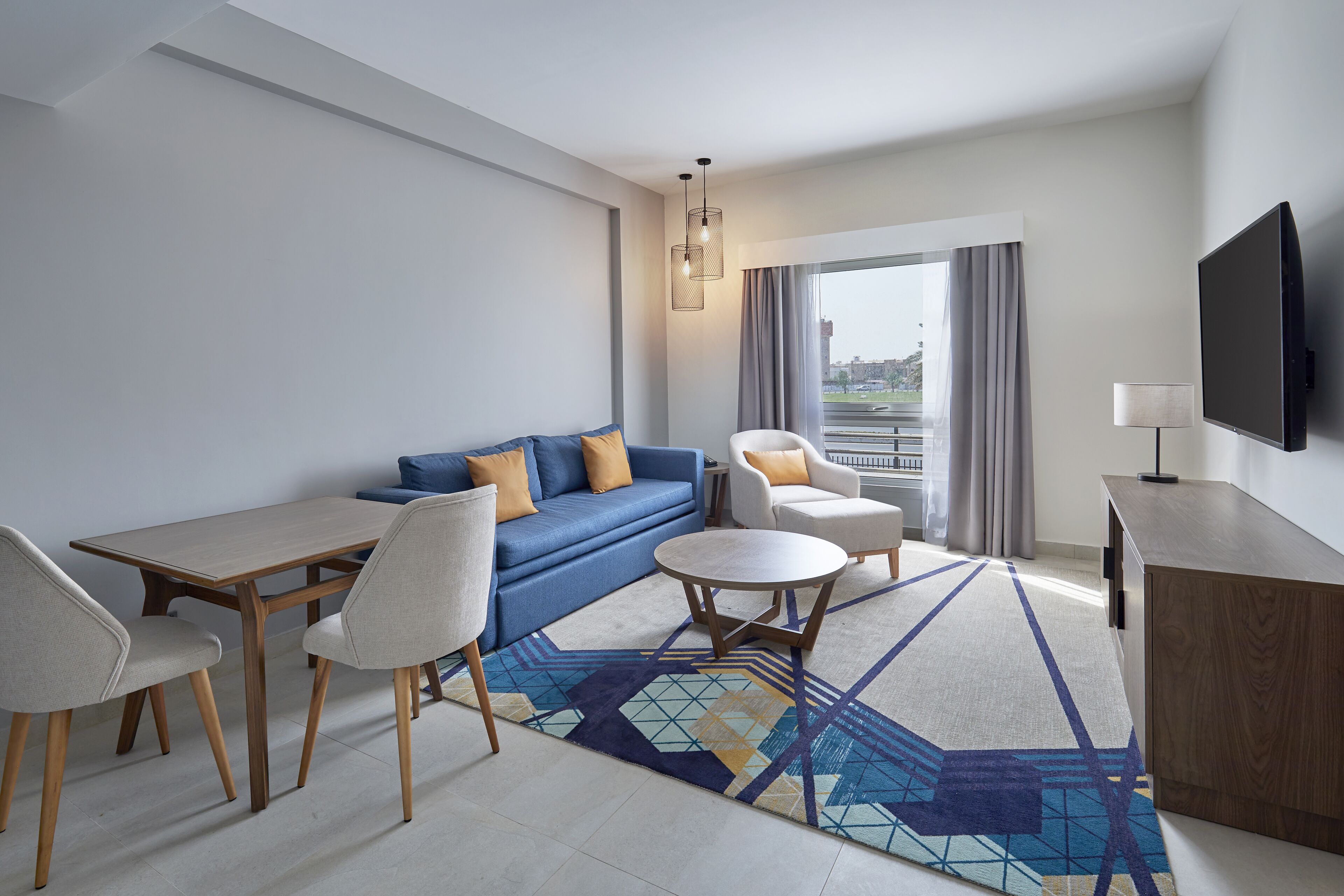 Una habitación de hotel moderna con sofá, sillas, una mesa de comedor, televisor y un amplio ventanal con cortinas.