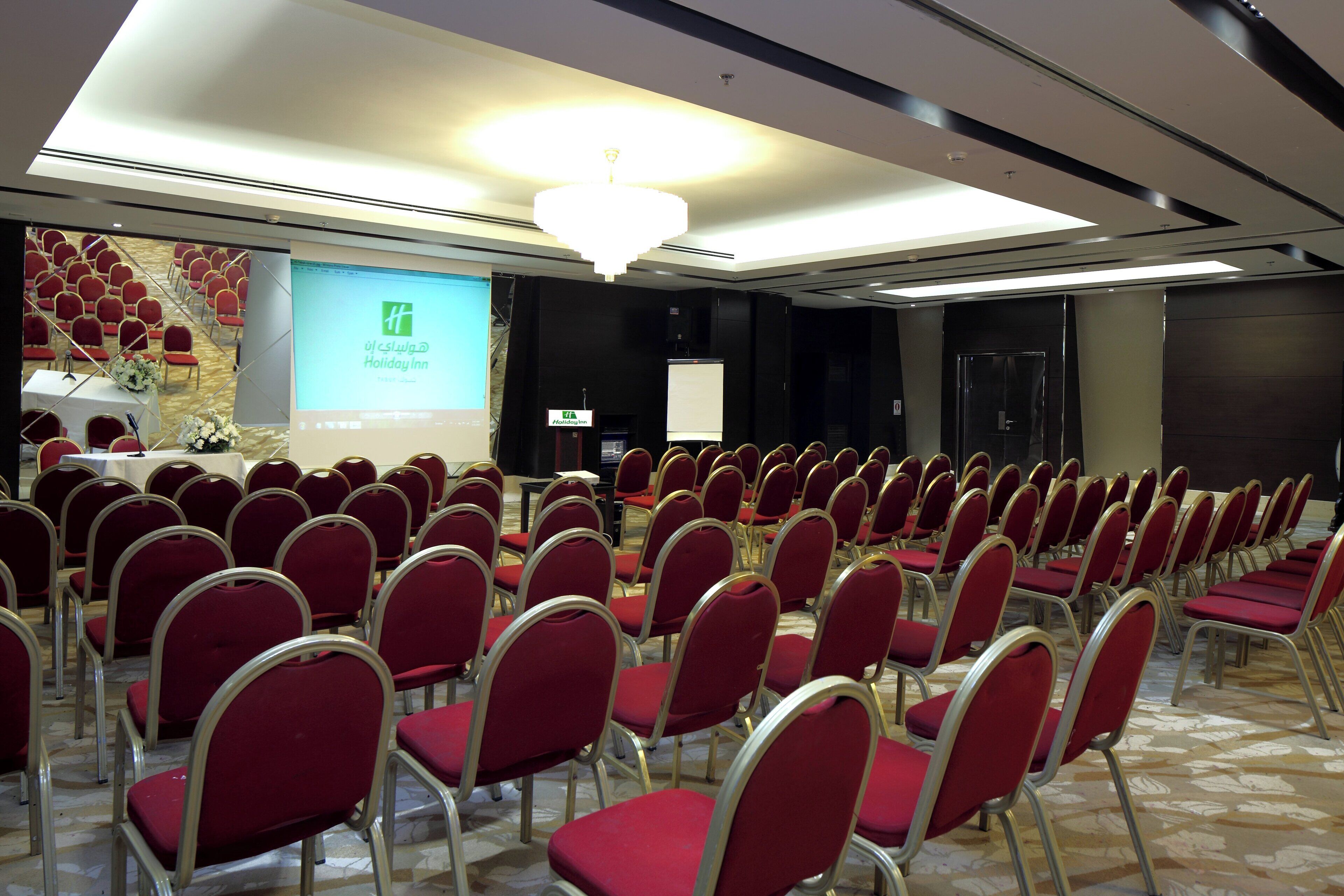 A conference room with rows of red chairs, a projection screen, and a stage with a table and microphone.
