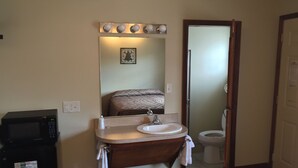 A bathroom with a sink, mirror, and toilet. There is a microwave on the left and a towel rack above the sink.