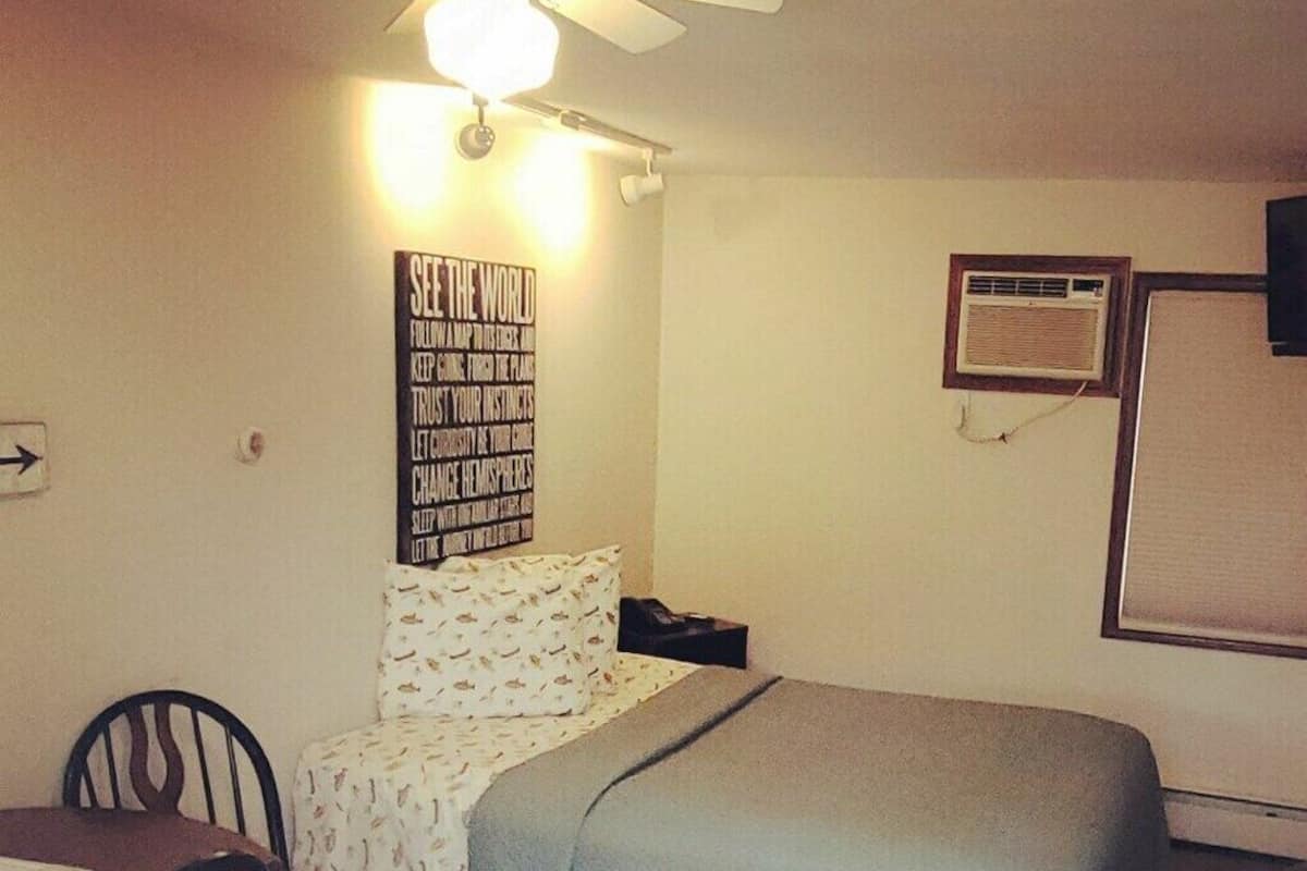 A small, neatly arranged bedroom with a bed, a desk, and a chair. There is a ceiling fan, a wall-mounted air conditioner, and a motivational poster on the wall.