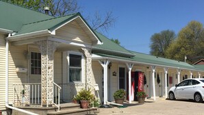 A row of single-story buildings with green roofs and white trim, each with a porch and potted plants.