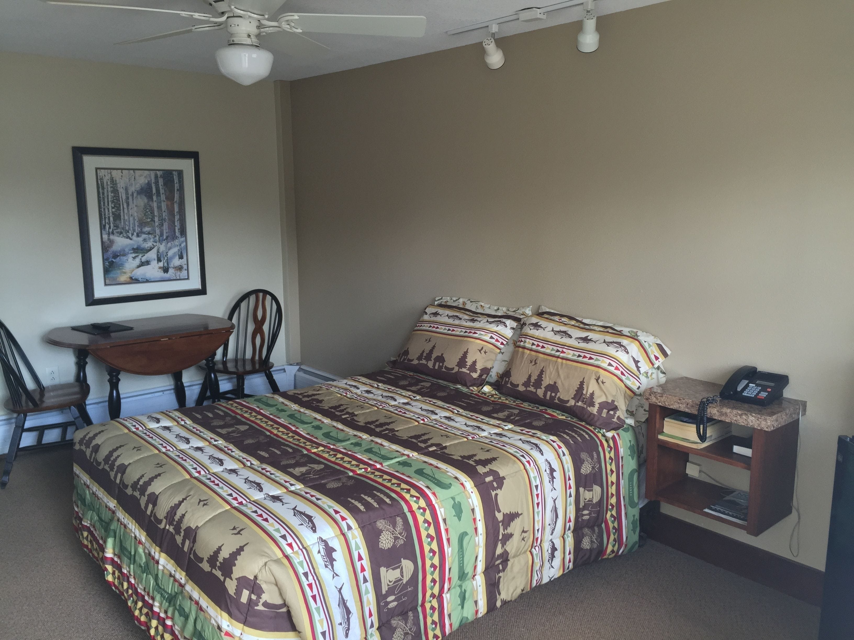 A bedroom with a bed, a desk, two chairs, a ceiling fan, and a telephone on a small table.