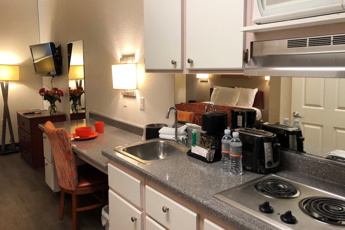 A compact hotel kitchen with a sink, stove, microwave, and a small dining area with a chair and table.