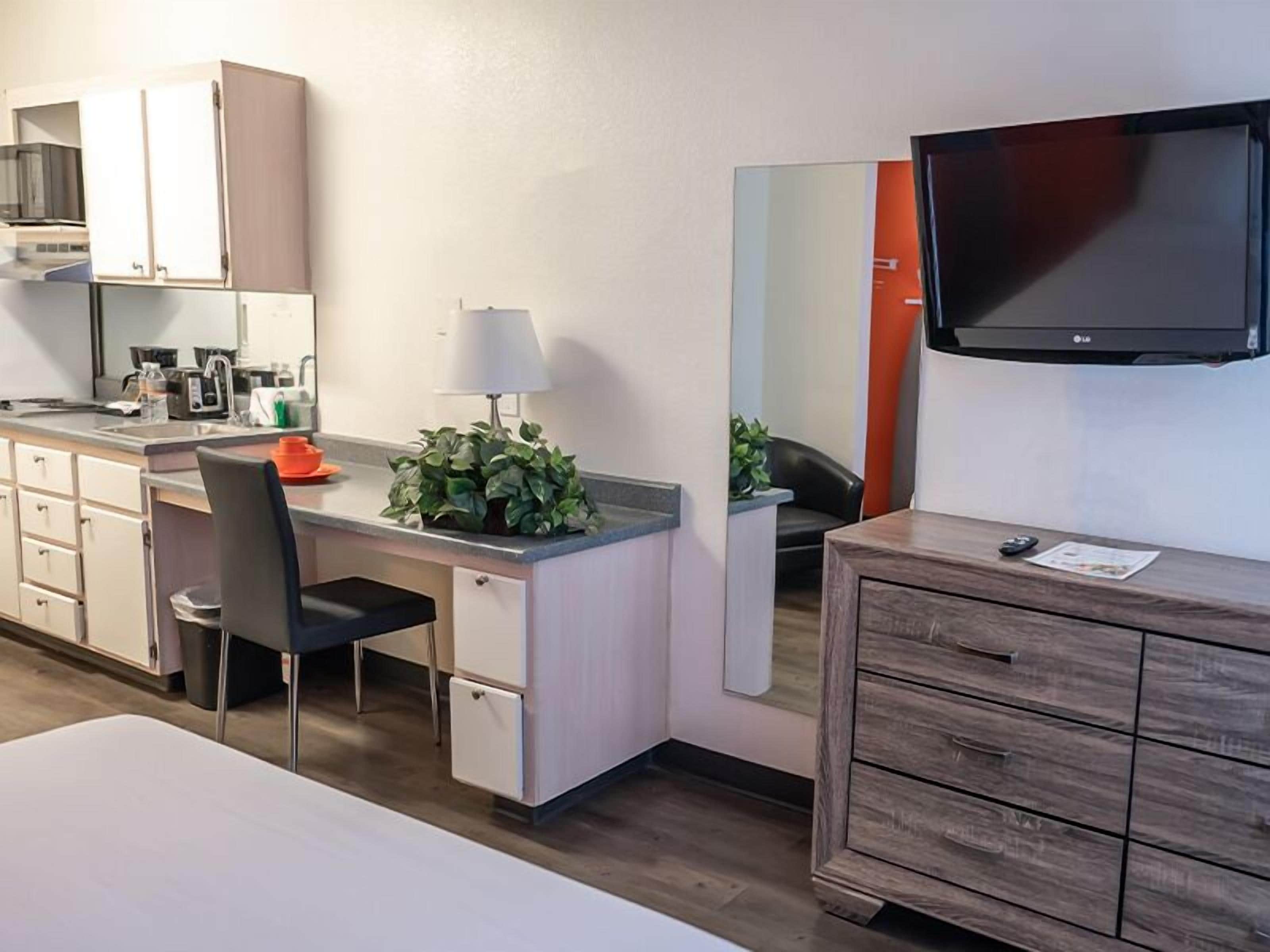 Habitación de hotel con kitchenette, escritorio con silla, pequeño armario, espejo, televisor y cómoda de madera.