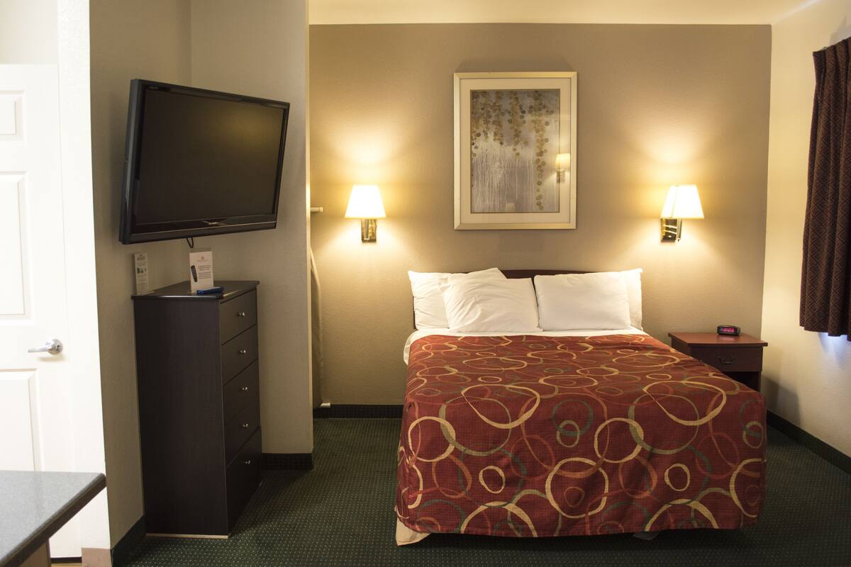 A hotel room with a bed, a television on a dresser, two wall-mounted lamps, and a painting on the wall.
