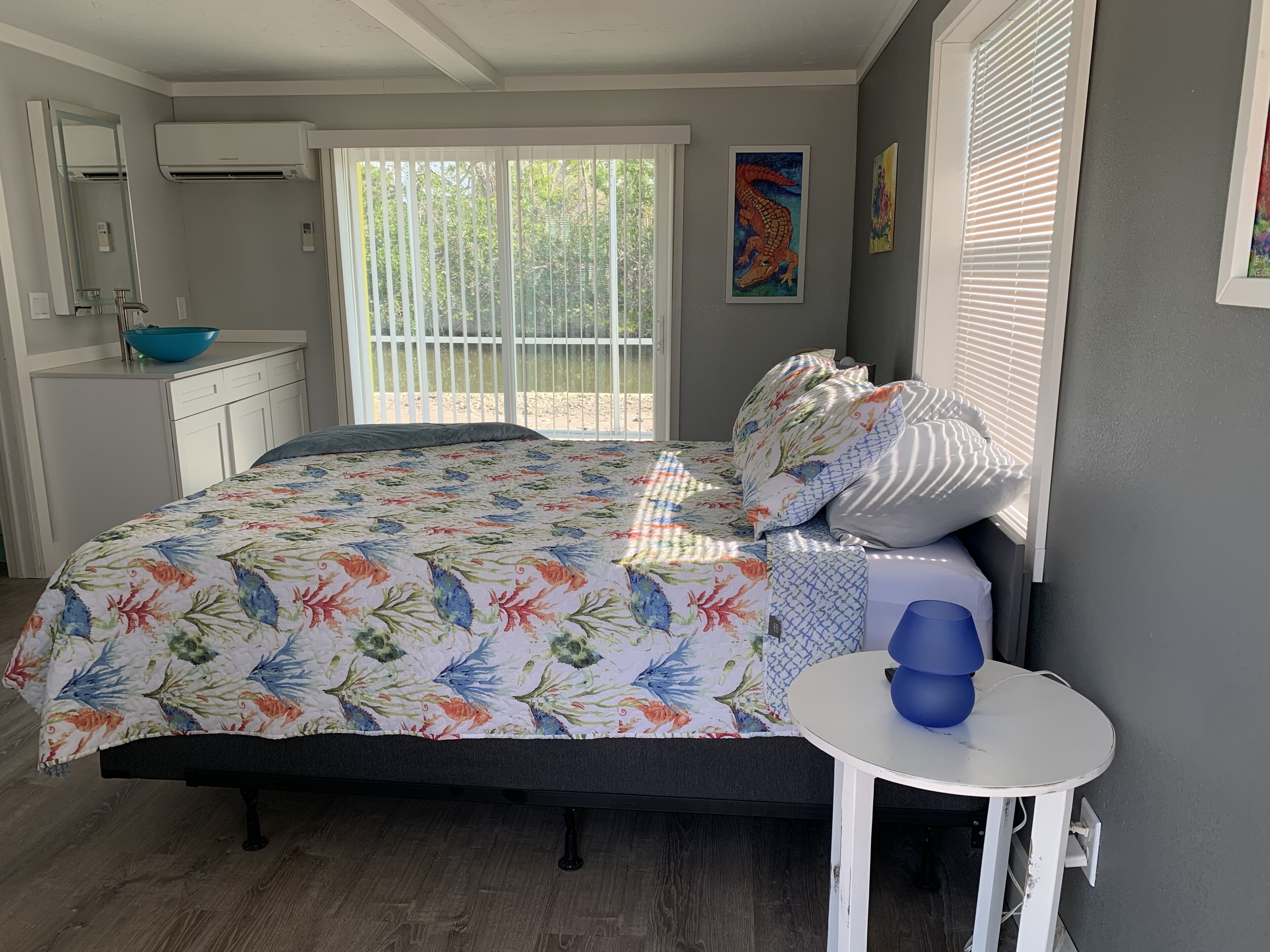 A bedroom with a bed, a small table, a window with blinds, a bathroom with a mirror and sink, and a door.