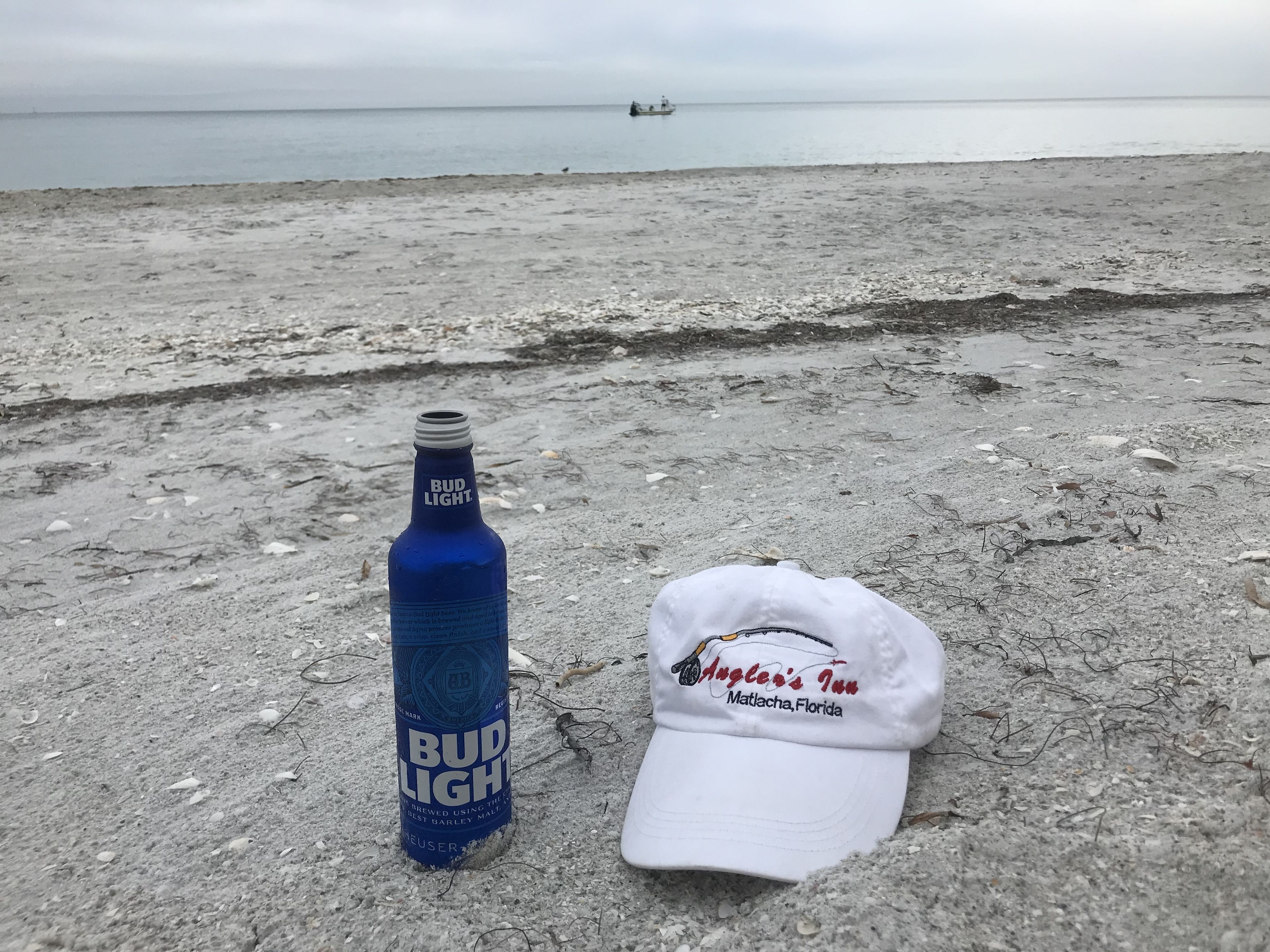 Bud Light -oluttölkki ja valkoinen lippalakko hiekkarannalla, taustalla näkyy vene.