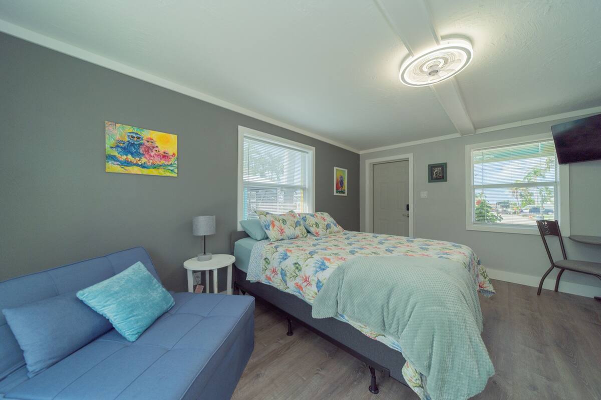 A bedroom with a bed, a blue sofa, a small table, and a chair. There are two windows with blinds, a ceiling light, and a painting on the wall.