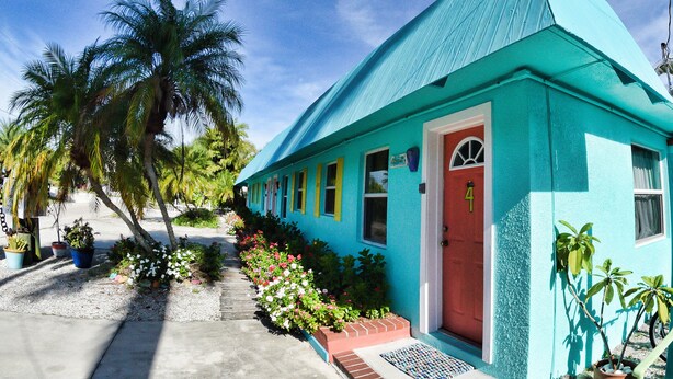 A turquoise building with a red door, surrounded by potted plants and flowers. - Angler's Inn (Matlacha)