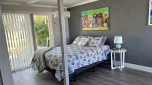 A bedroom with a bed, a nightstand, a painting, and a window with blinds. - Angler's Inn (Matlacha)