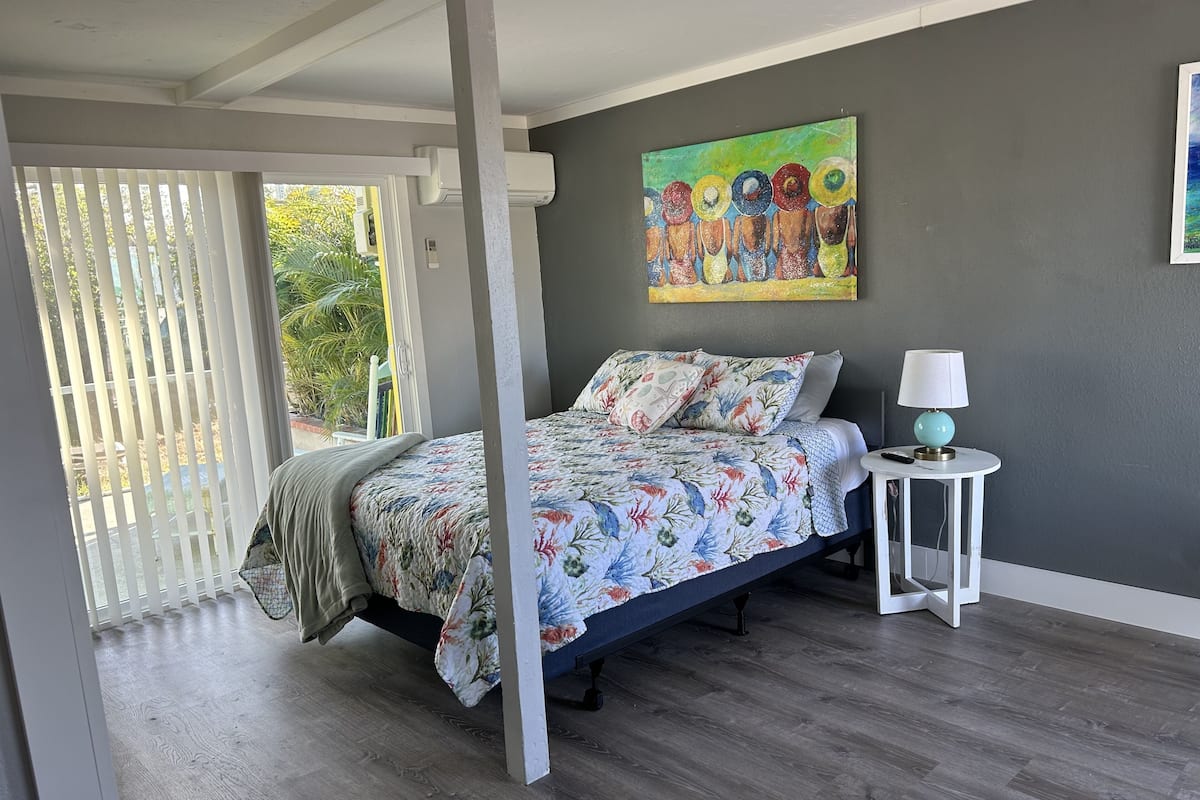 A bedroom with a bed, a nightstand, a painting, and a window with blinds.