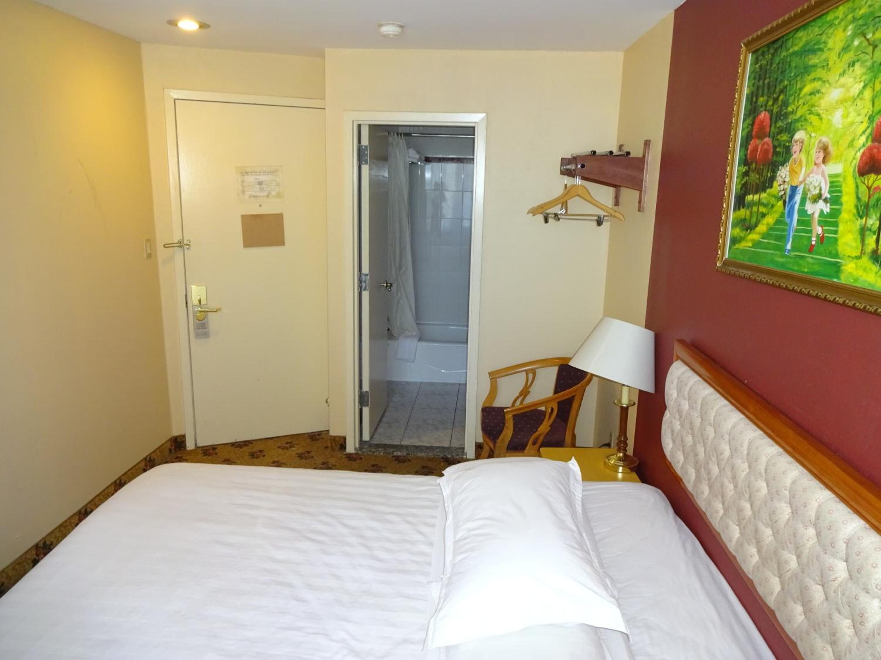 A hotel room with a bed, a door, a bathroom, a chair, a lamp, and a painting on the wall.