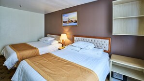 A hotel room with two beds, a nightstand with a lamp, a framed cityscape picture on the wall, and a wooden shelf unit. - Flushing Hotel (Flushing)