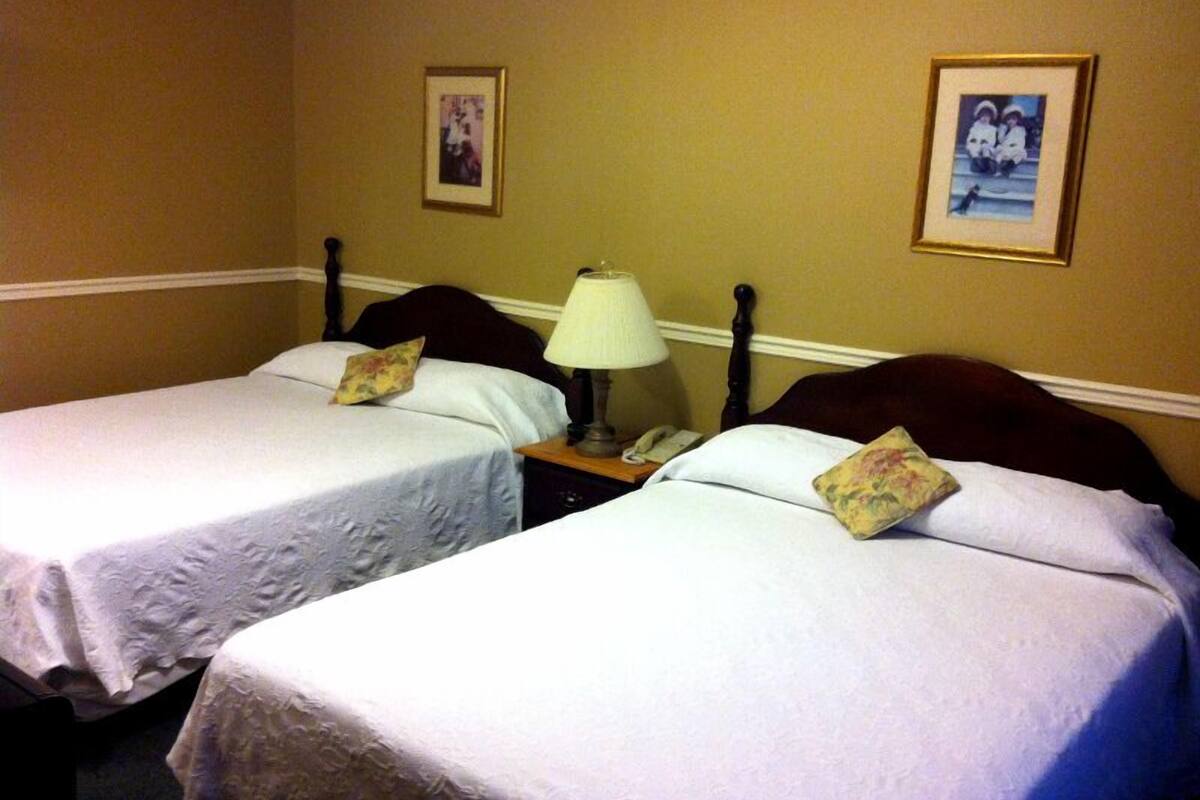 Habitación de hotel con dos camas individuales, una mesita de noche con lámpara y cuadros en las paredes.