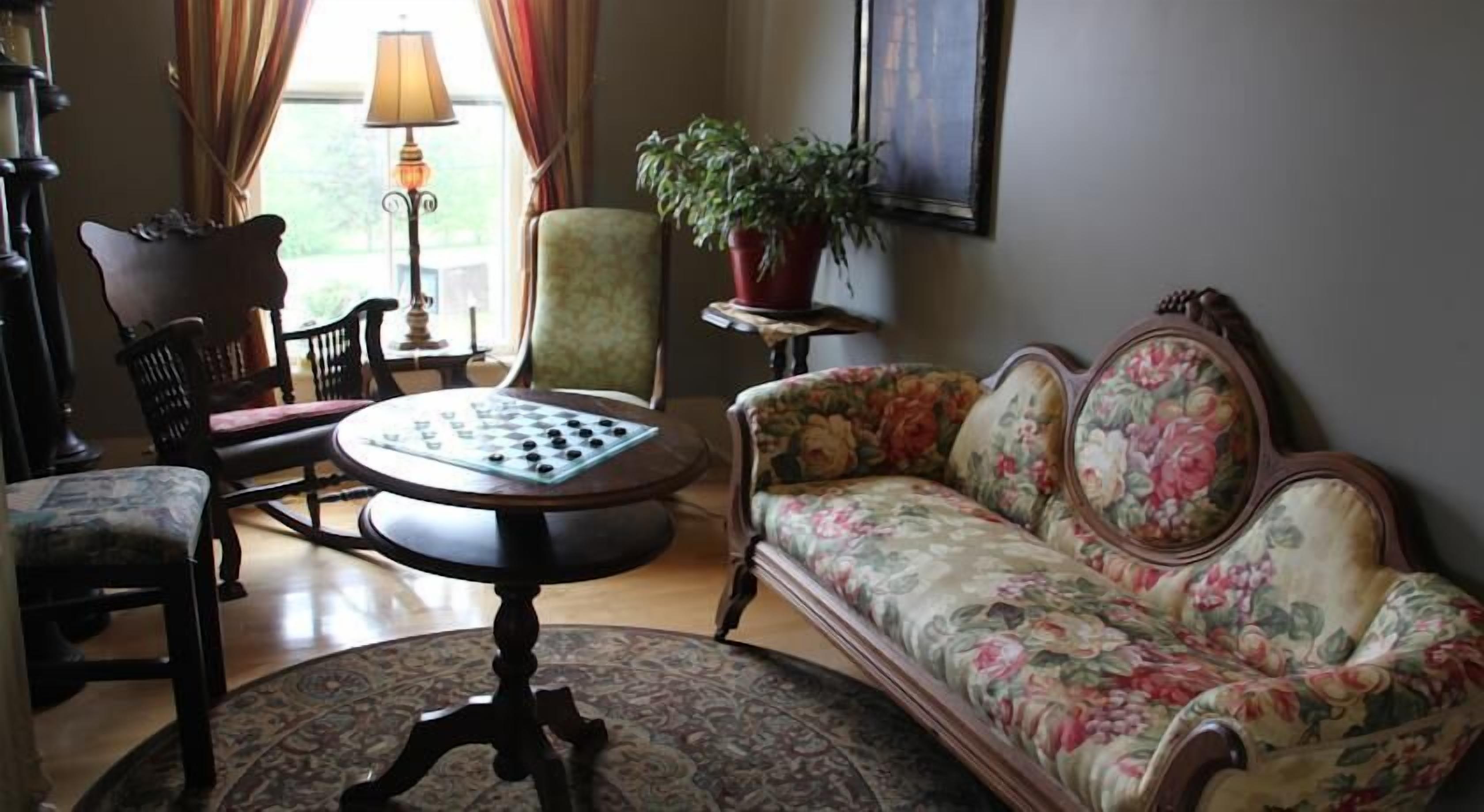 Una sala de estar con un sofá floral, una mesa redonda con un tablero de ajedrez, una planta en maceta y una lámpara.