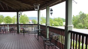 A covered outdoor area with wooden flooring, metal chairs, and tables, featuring a view of trees and a distant mountain. - Lynwood Inn (Baddeck)
