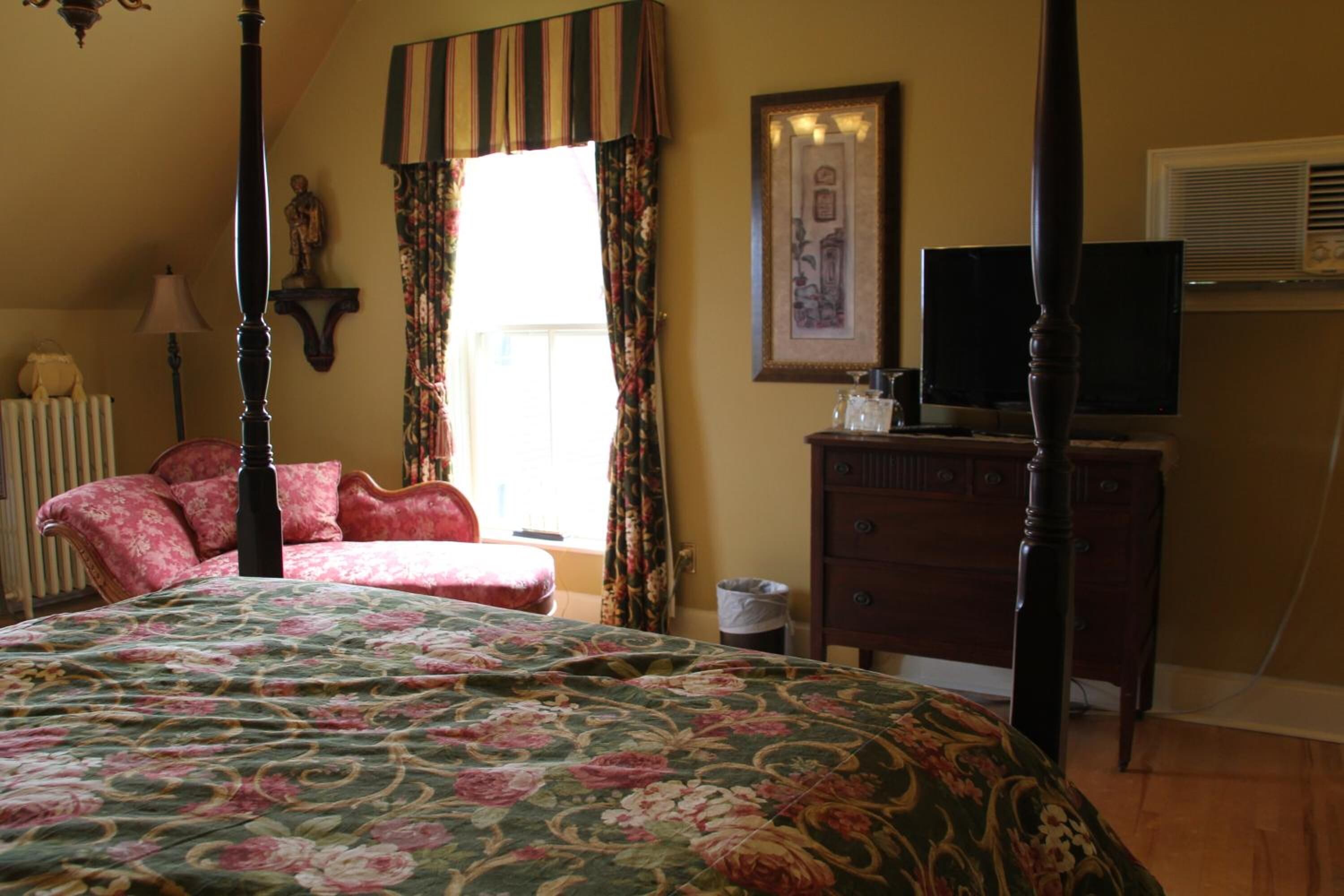 A bedroom with a four-poster bed, a dresser, a television, and a window with curtains.