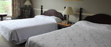 Habitación de hotel con dos camas individuales, una mesita de noche con lámpara, una silla y un cuadro en la pared.