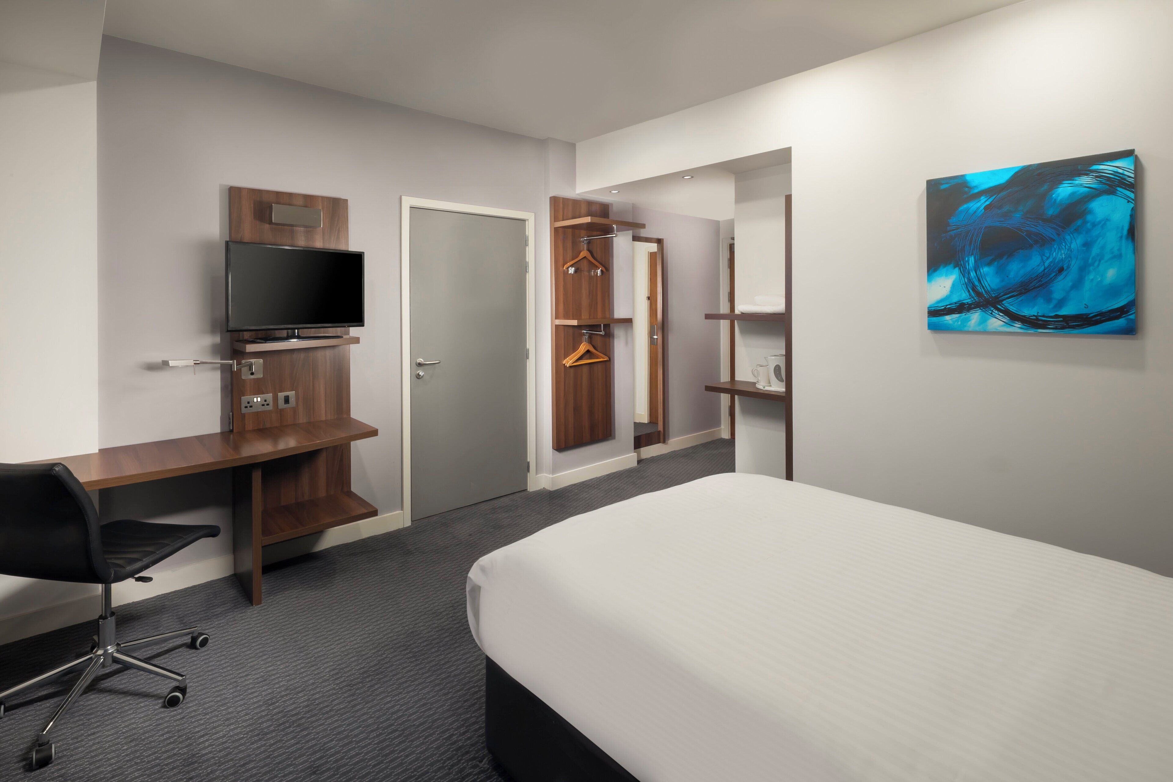 A hotel room with a bed, a desk with a TV, a wardrobe, and a painting on the wall.