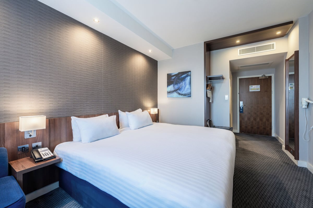 A hotel room with a large bed, a desk with a telephone, a wall-mounted lamp, and a door leading to a bathroom.