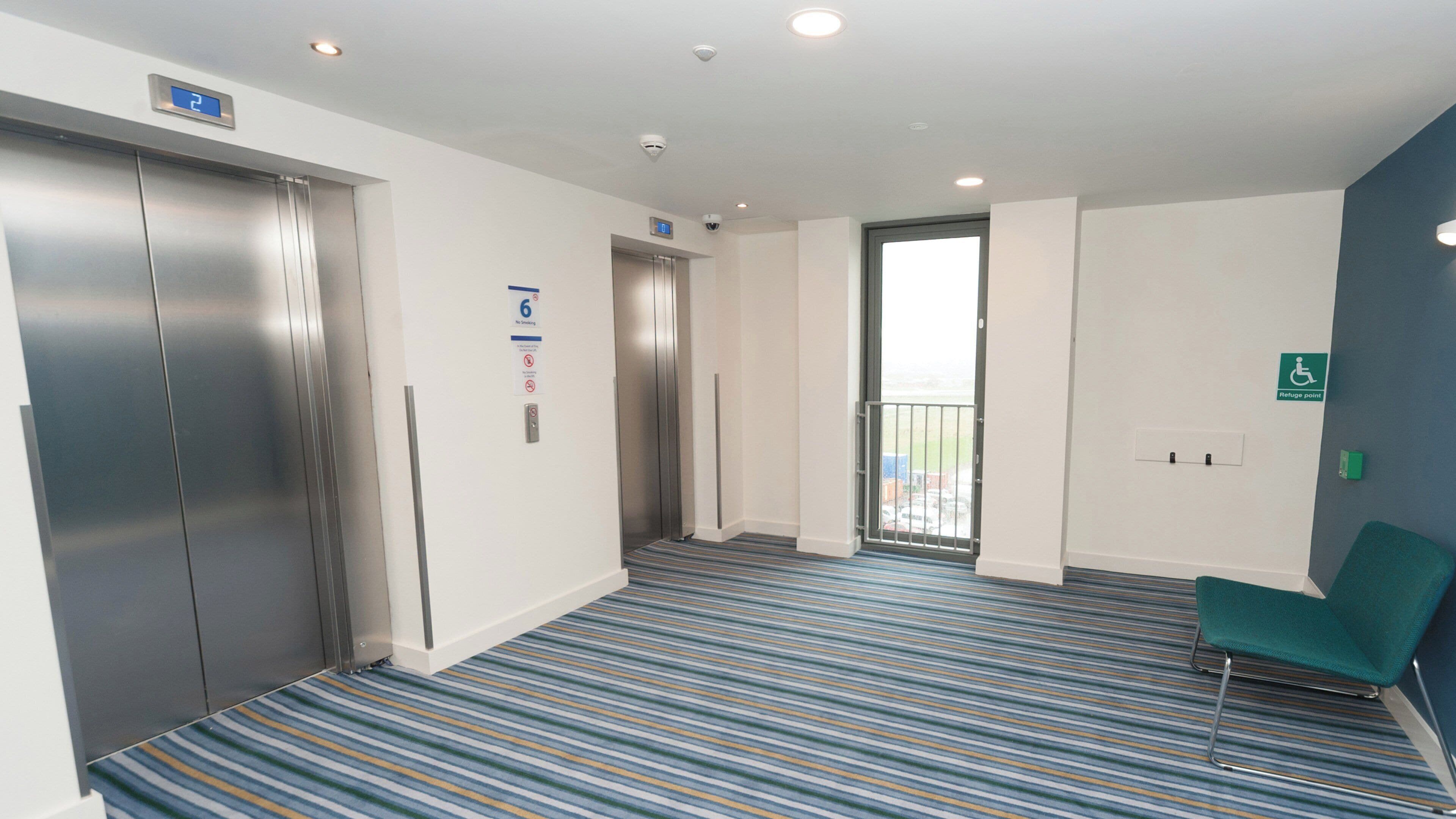 Et hotel lobby med et stribet tæppe, to elevatorer, en grøn stol og et skilt, der angiver adgang for kørestolsbrugere.