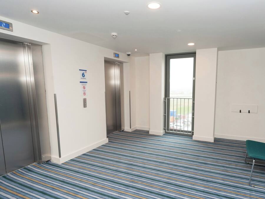 Et hotel lobby med et stribet tæppe, to elevatorer, en grøn stol og et skilt, der angiver adgang for kørestolsbrugere.