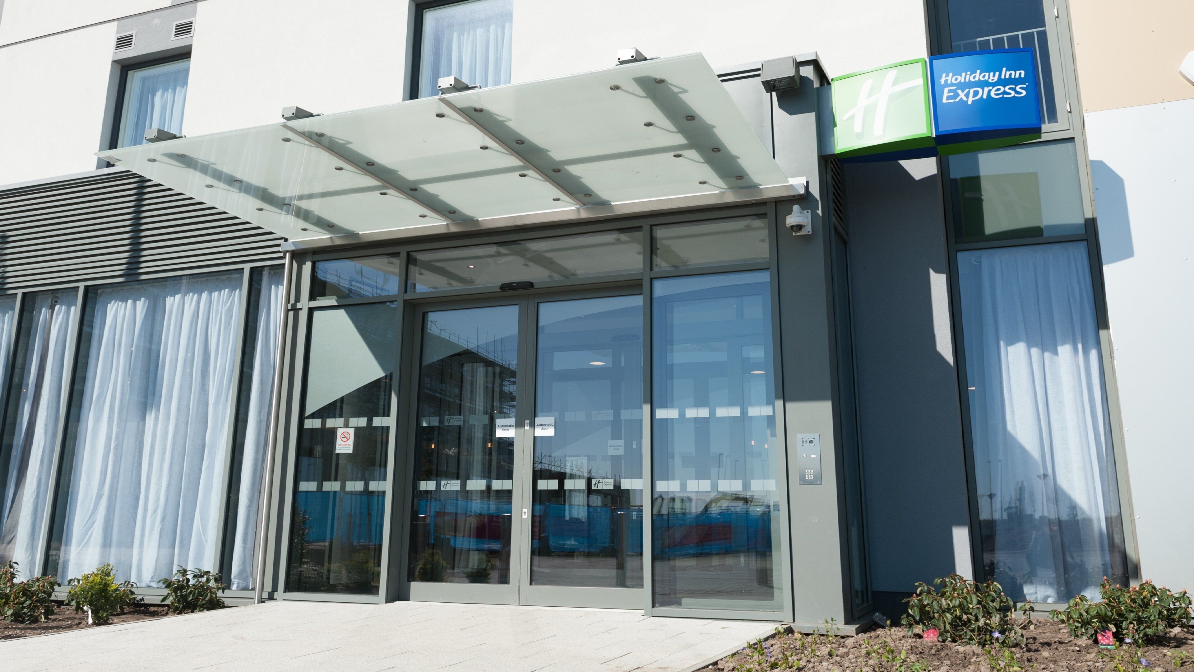 A Holiday Inn Express building with a glass entrance, a covered waiting area, and a sign indicating the hotel name.