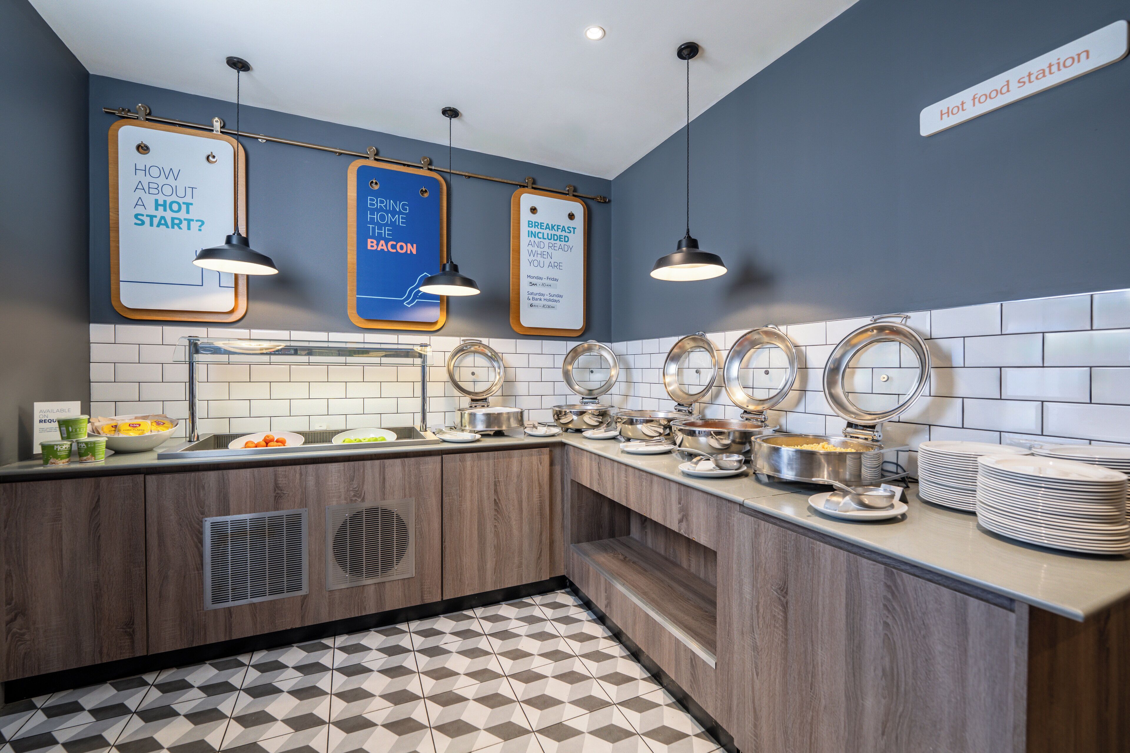 Un buffet avec plusieurs points de service, un panneau indiquant que le petit-déjeuner est inclus, et un autre panneau annonçant l’ouverture d’un espace où des plats chauds seront servis.