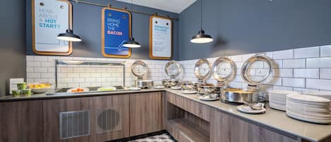Un buffet avec plusieurs points de service, un panneau indiquant que le petit-déjeuner est inclus, et un autre panneau annonçant l’ouverture d’un espace où des plats chauds seront servis.