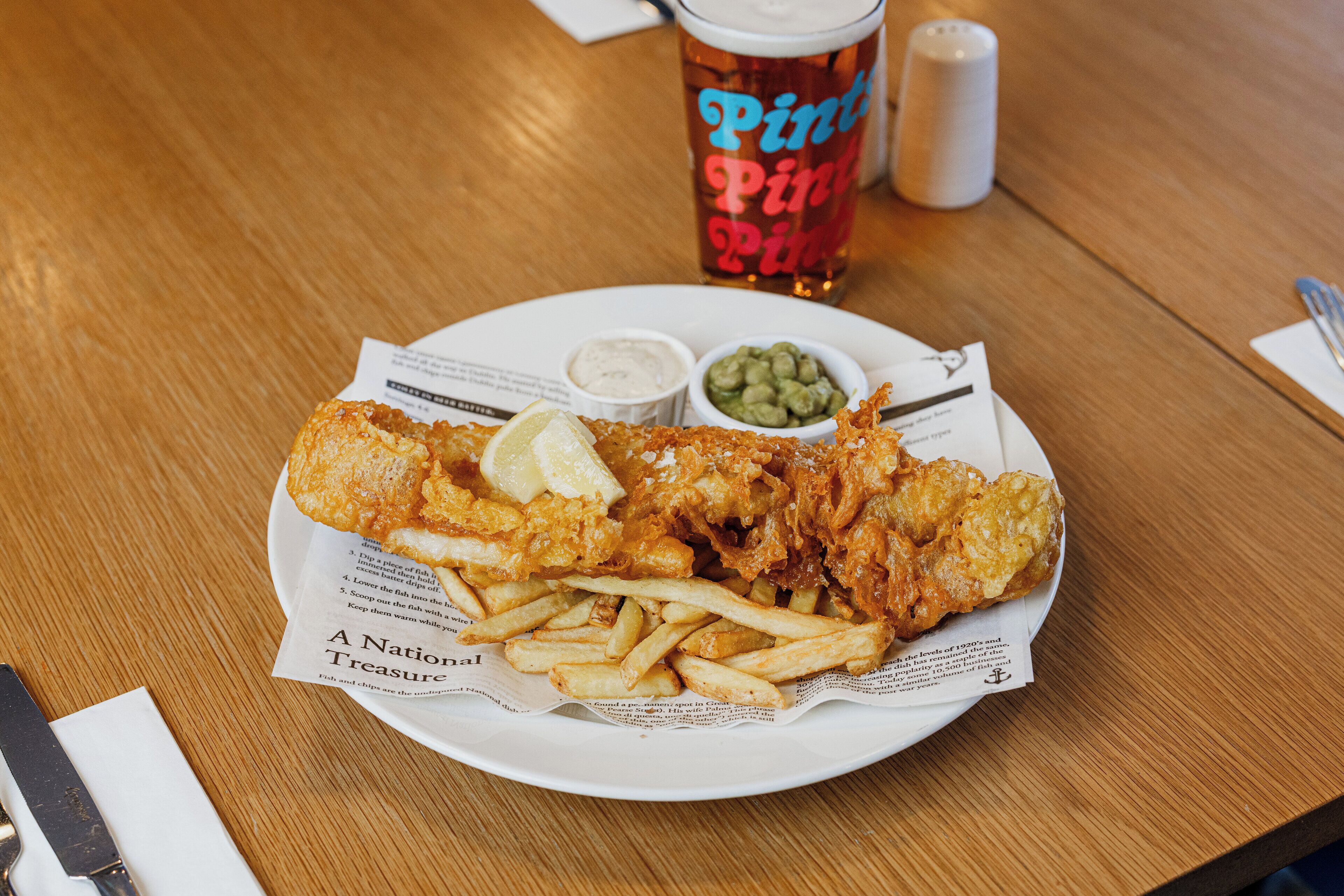 Un plato de pescado con papas fritas, una bebida y salsas en una mesa de madera.