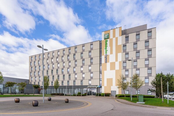 A modern hotel building with a checkered facade, a prominent green and white sign, and a landscaped courtyard with bollards and trees.